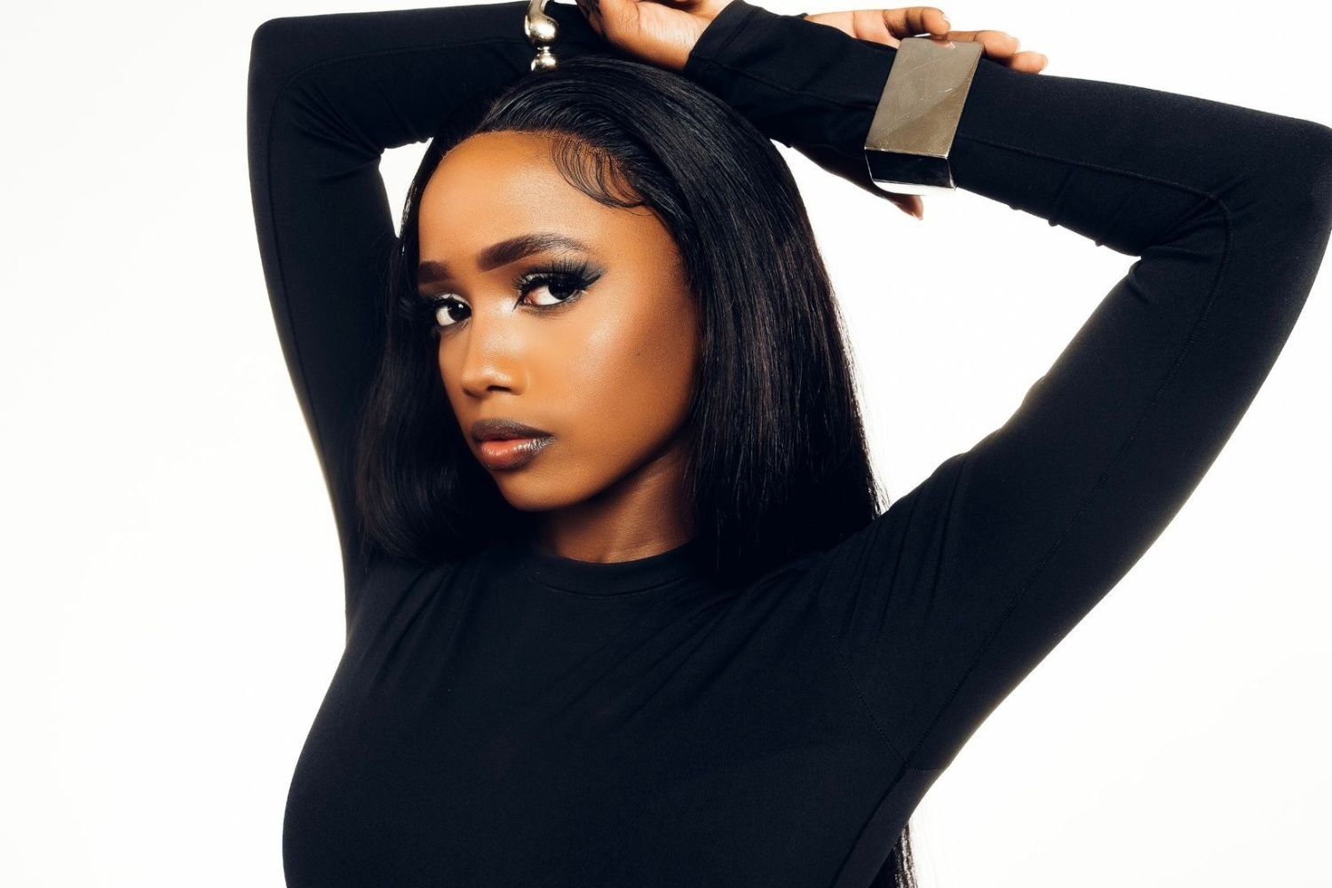 Abigail Chams, a woman with sleek, straight hair and flawless makeup poses confidently with her arms raised above her head, wearing a fitted black top and a chunky metallic bracelet, set against a plain white background.