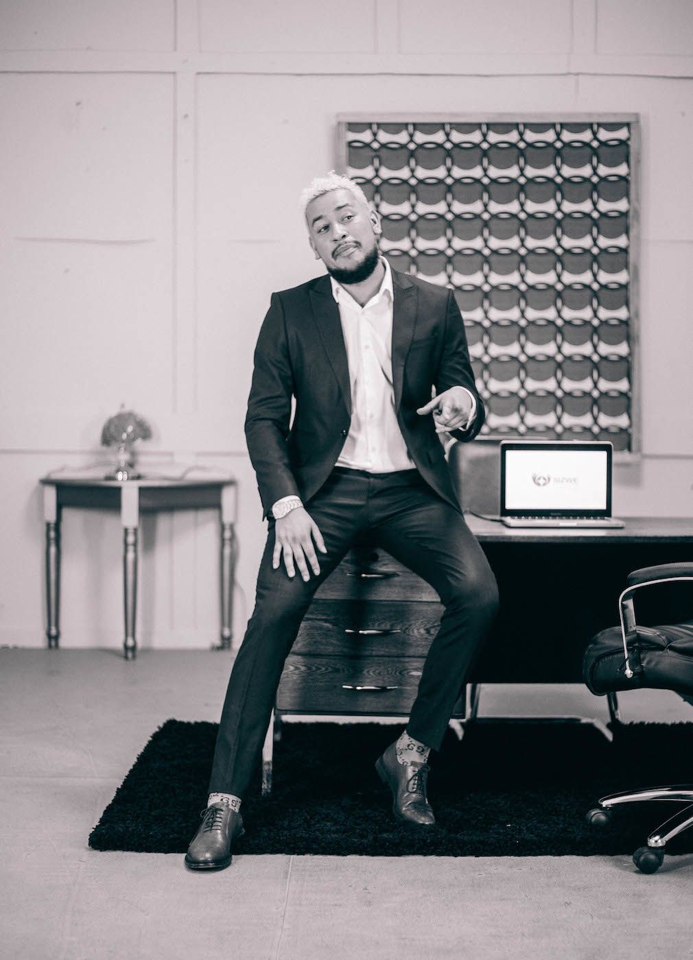 A portrait of AKA in a suit sitting on an office desk. 