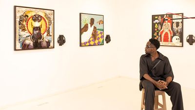 An image of Mr Eazi sitting on a chair in a gallery, looking back at three artworks hanging on the wall. 