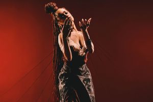 A press shot of Nanette against a red background. She is dressed in a black dress with a slit, wears black boots, and holds strings with her fingers. 