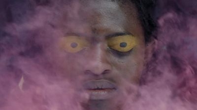 A still from Baloji's Omen of a pair of eyes with yellow eyeshadow on them covered by a cloud of pink smoke.
