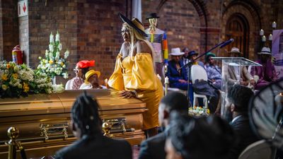 A still from the TV series, of a woman dressed in yellow standing at a coffin.