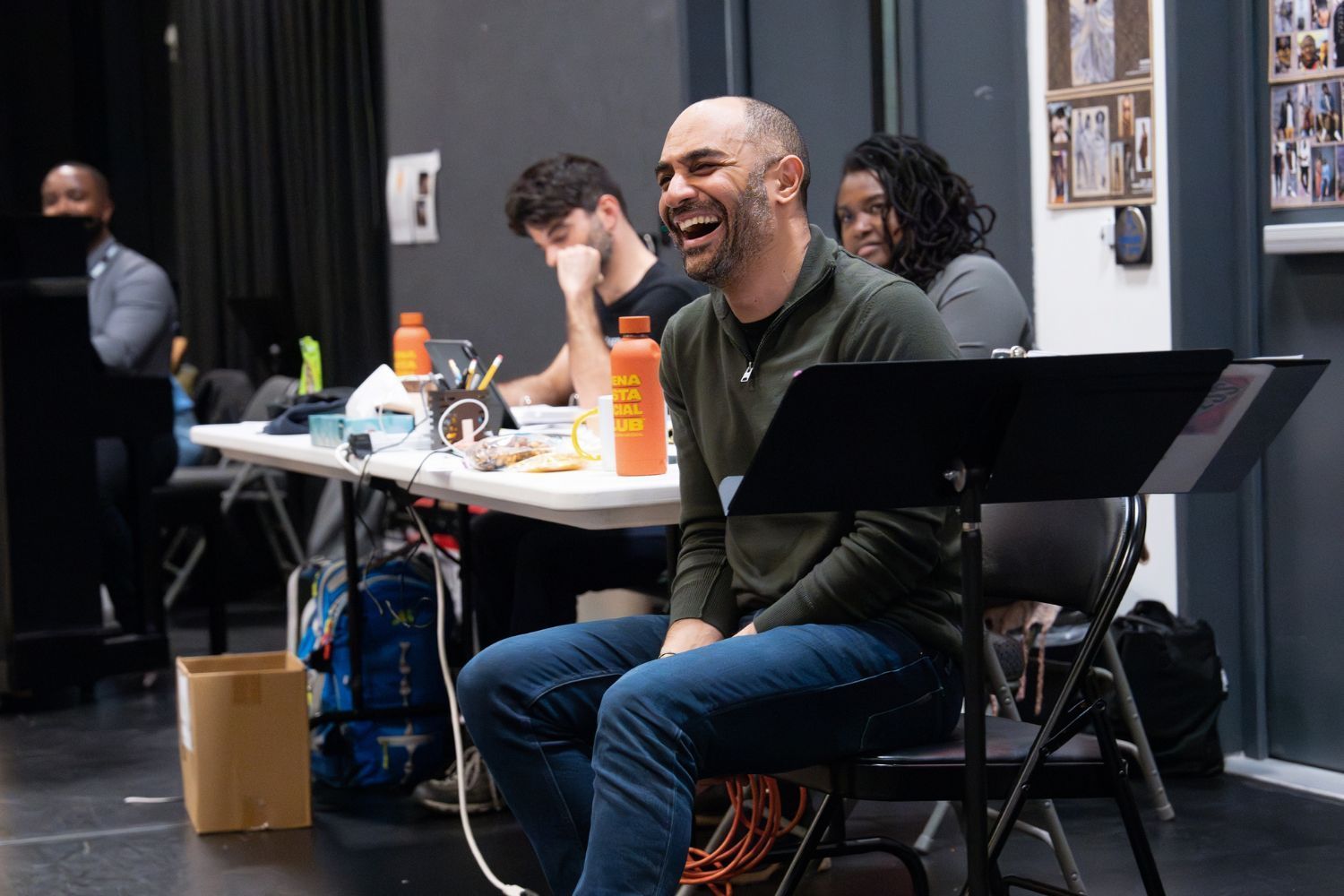Saheem Ali laughing during a rehearsal, seated in front of a table with scripts and snacks, surrounded by collaborators at the Public Theater.