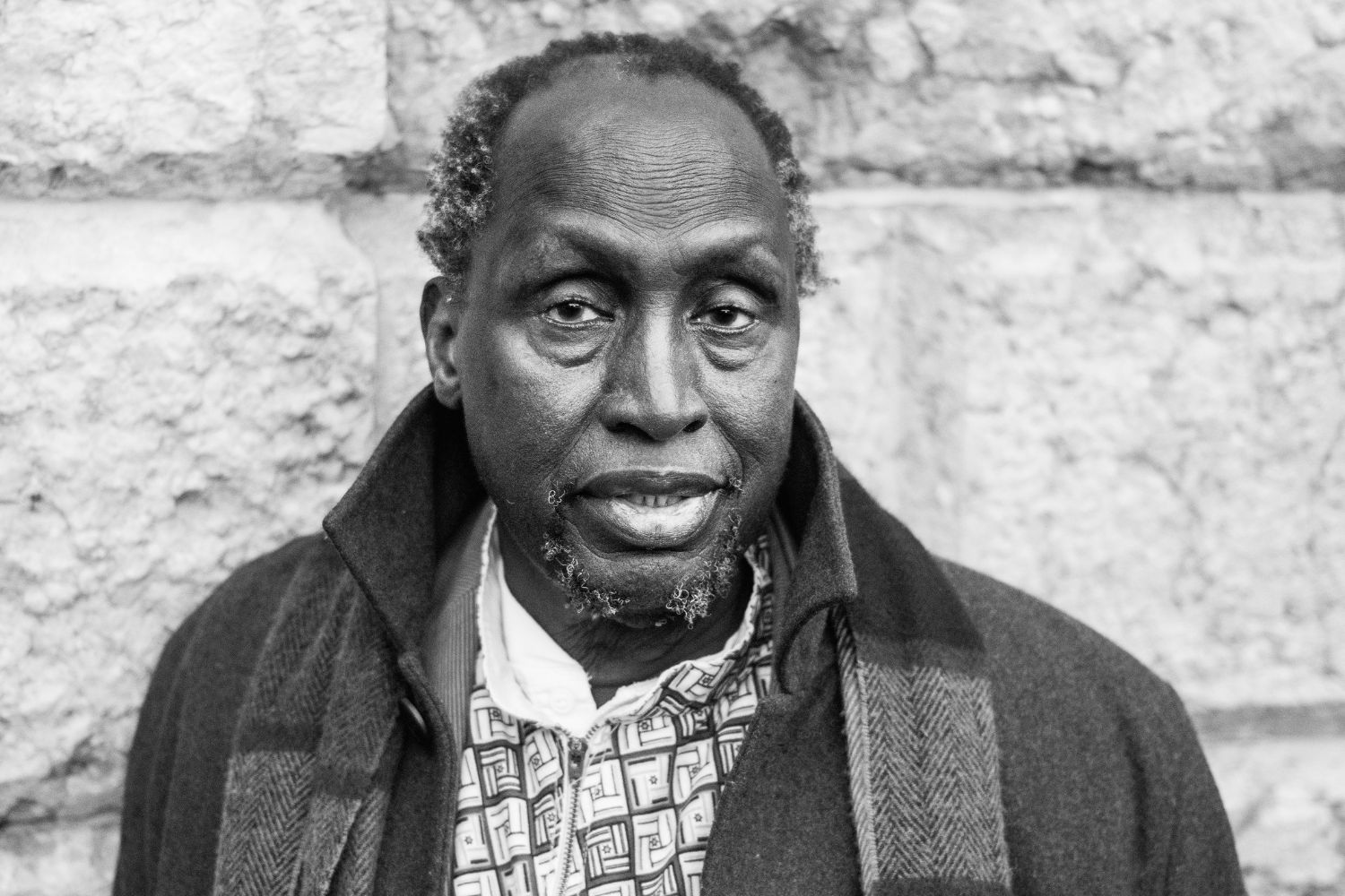 Late Kenyan author Ngũgĩ wa Thiong’o stands in front of a stone wall, wearing a patterned shirt and a dark coat with a scarf draped over his shoulders.