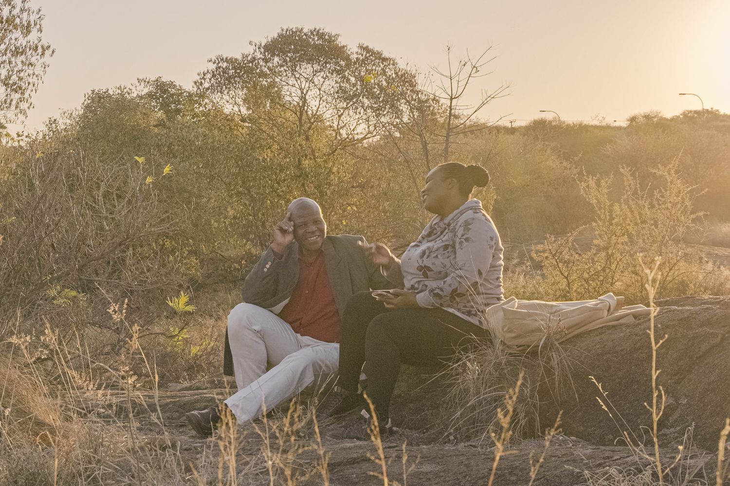 Chris and Dumi sit on a rock in a sunlit field in Botswana, sharing a quiet moment in a scene from Matabeleland, directed by Nyasha Kadandara.