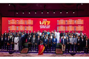 A group of international football delegates stands on stage in front of a red backdrop displaying the official group draw for the FIFA U-17 World Cup Qatar 2025. The background shows the tournament logo and twelve groups labeled A through L, each listing four competing nations. At the center foreground, a trophy rests on a podium, with the tournament’s branding clearly visible. 