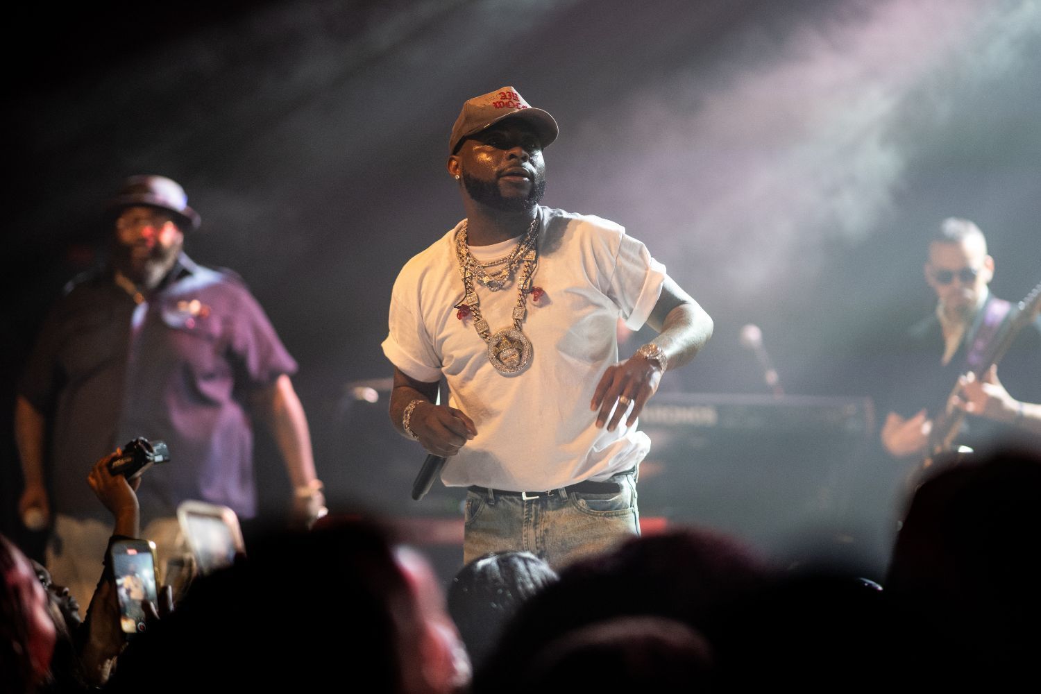Davido in a white t-shirt, cap, and heavy chains energizes a crowd during a live concert.