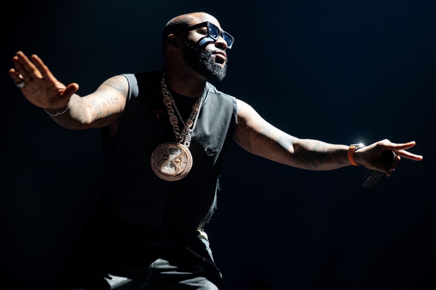 Davido wears a sleeveless black outfit and oversized chain and strikes a dynamic pose under a spotlight on a stage.