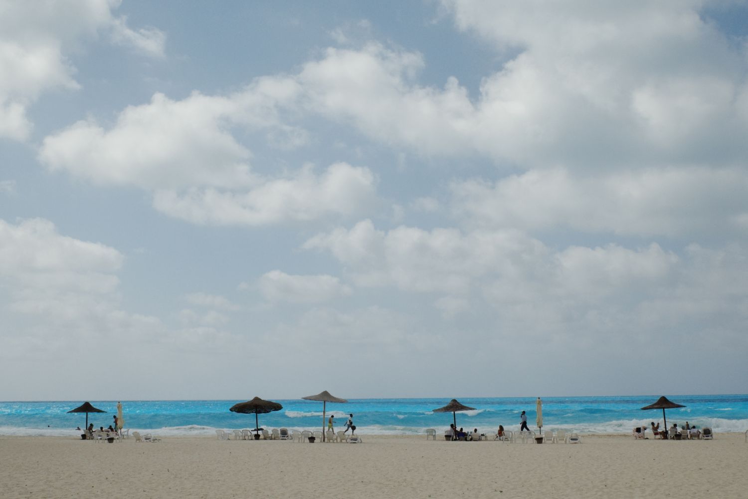 A Mediterranean shoreline with white umbrellas. Few people are walking on the beach. 