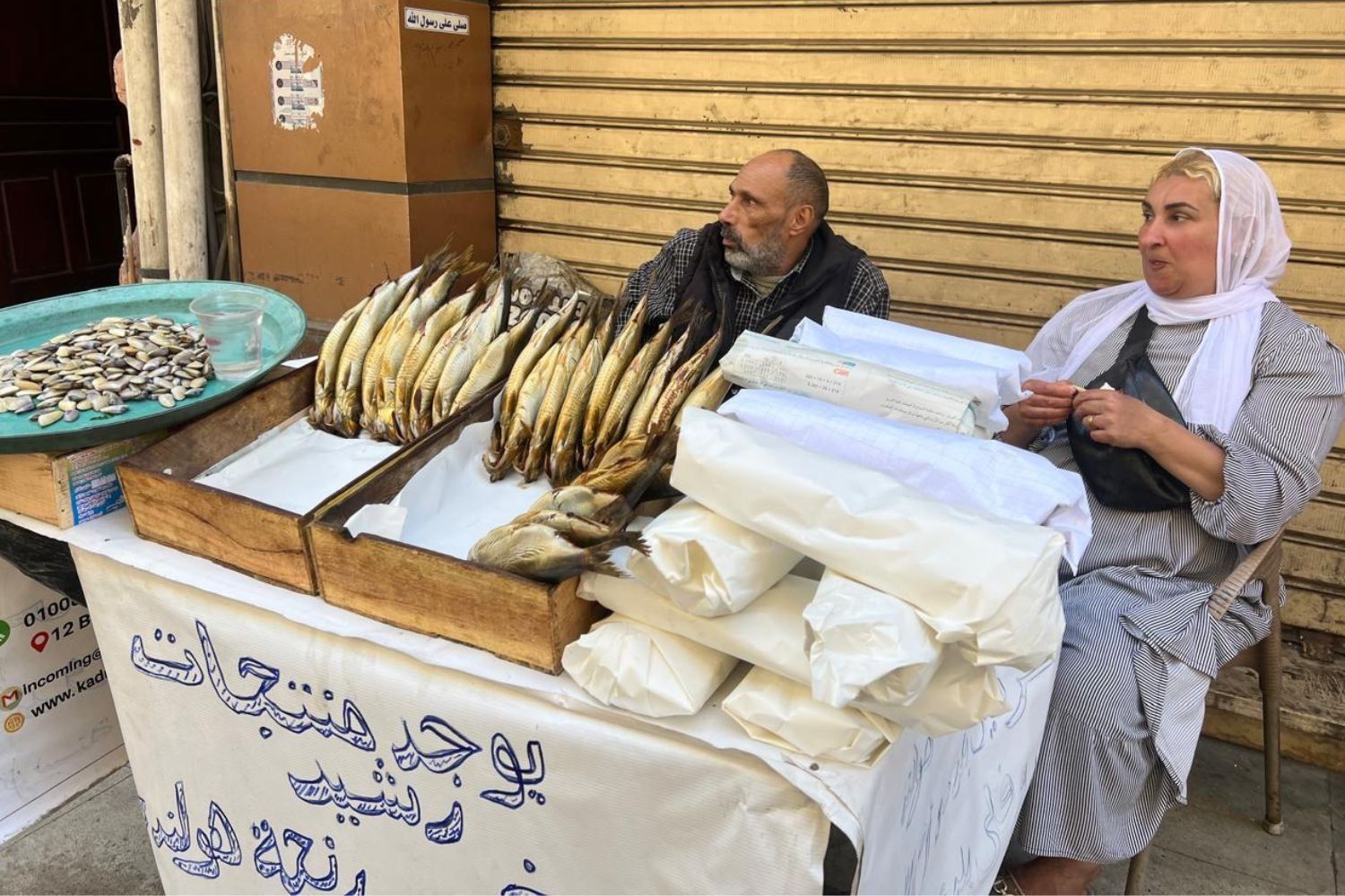 Samah Saeed and her brother Salah Abdelaziz are sitting behind a table that offers fermented, salted fish and mussels. They are wearing the traditional clothes of Egypt’s countryside. Blonde wavy hair is showing under Saeed’s white hijab. 