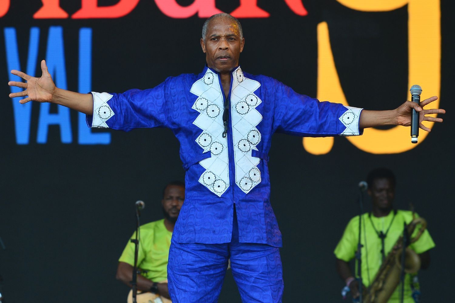 Femi Kuti, in a vibrant blue outfit, holds a microphone and spreads his arms wide on stage, with musicians playing in the background.