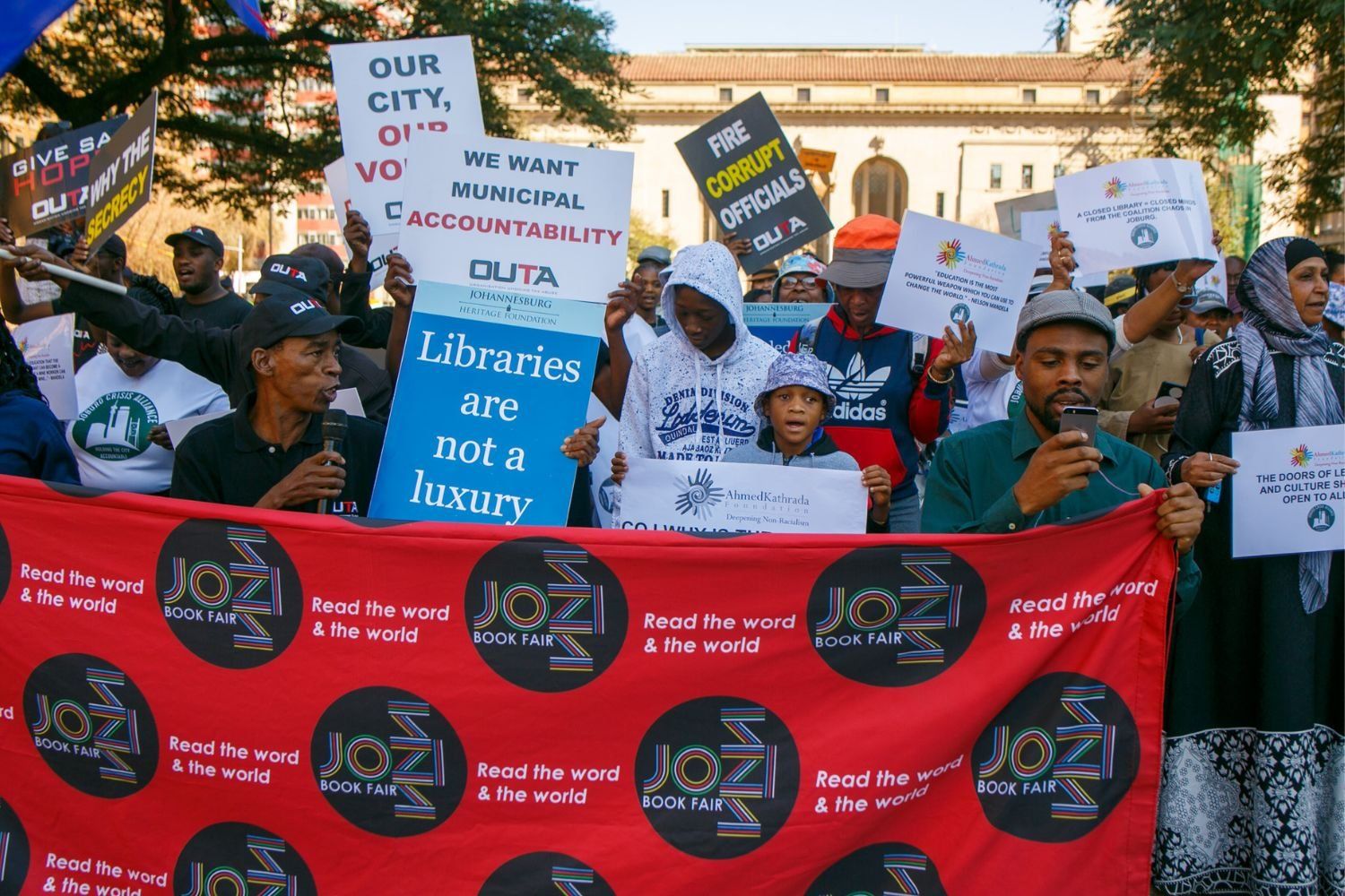 A group of people protest for public access to libraries in Johannesburg, South Africa. They hold signs reading “Libraries are not a luxury,” “Fire corrupt officials,” and “We want municipal accountability.” A red banner at the front reads “Jozi Book Fair: Read the word & the world.” The atmosphere is peaceful and determined.