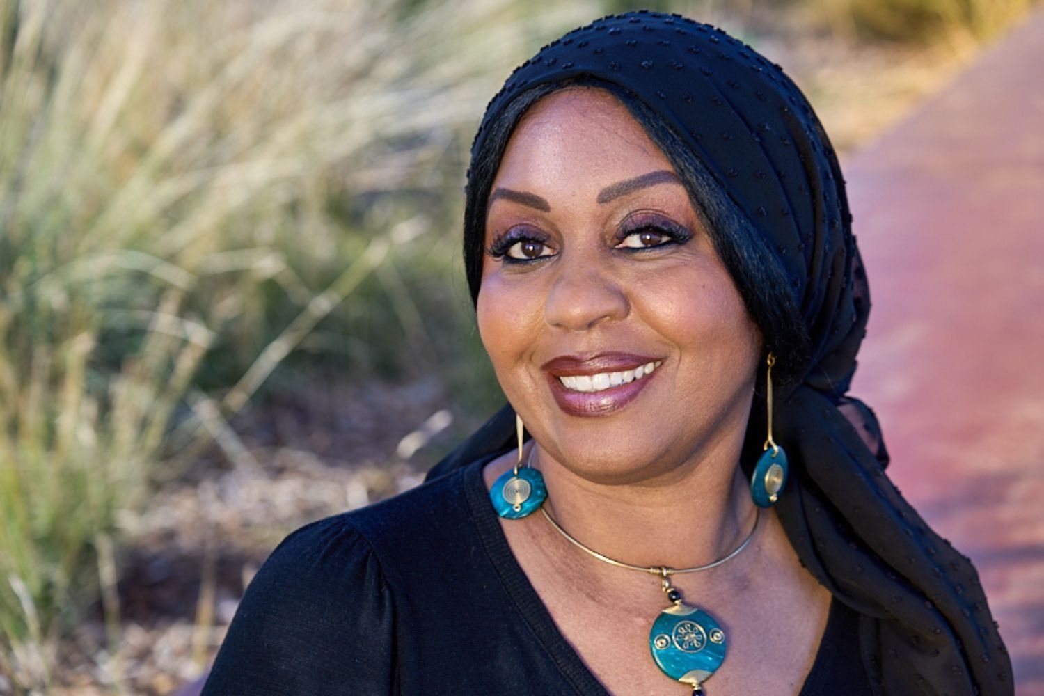 Portrait of Sudanese American radio journalist Hana Baba as she smiles into the camera, wearing a black hijab and blue and golden jewellery. 