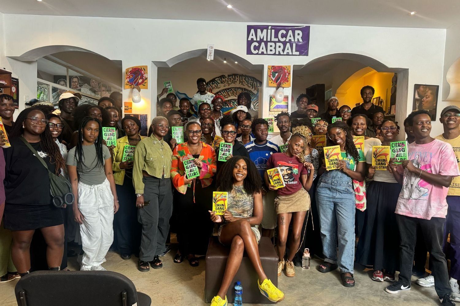 A large group of young people gathers indoors, many holding copies of the book Chain-Gang All-Stars, with one woman seated front and center under a banner that reads “Amílcar Cabral.”