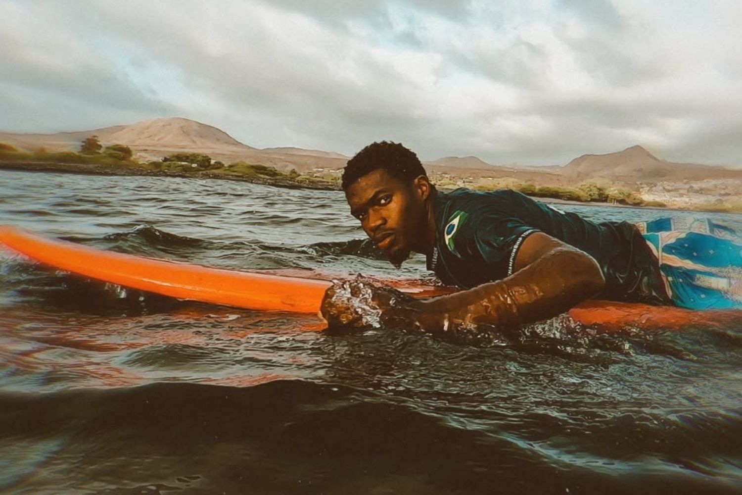 A man lies on a bright orange surfboard in the water, looking intensely toward the camera, with mountains and cloudy skies in the background.