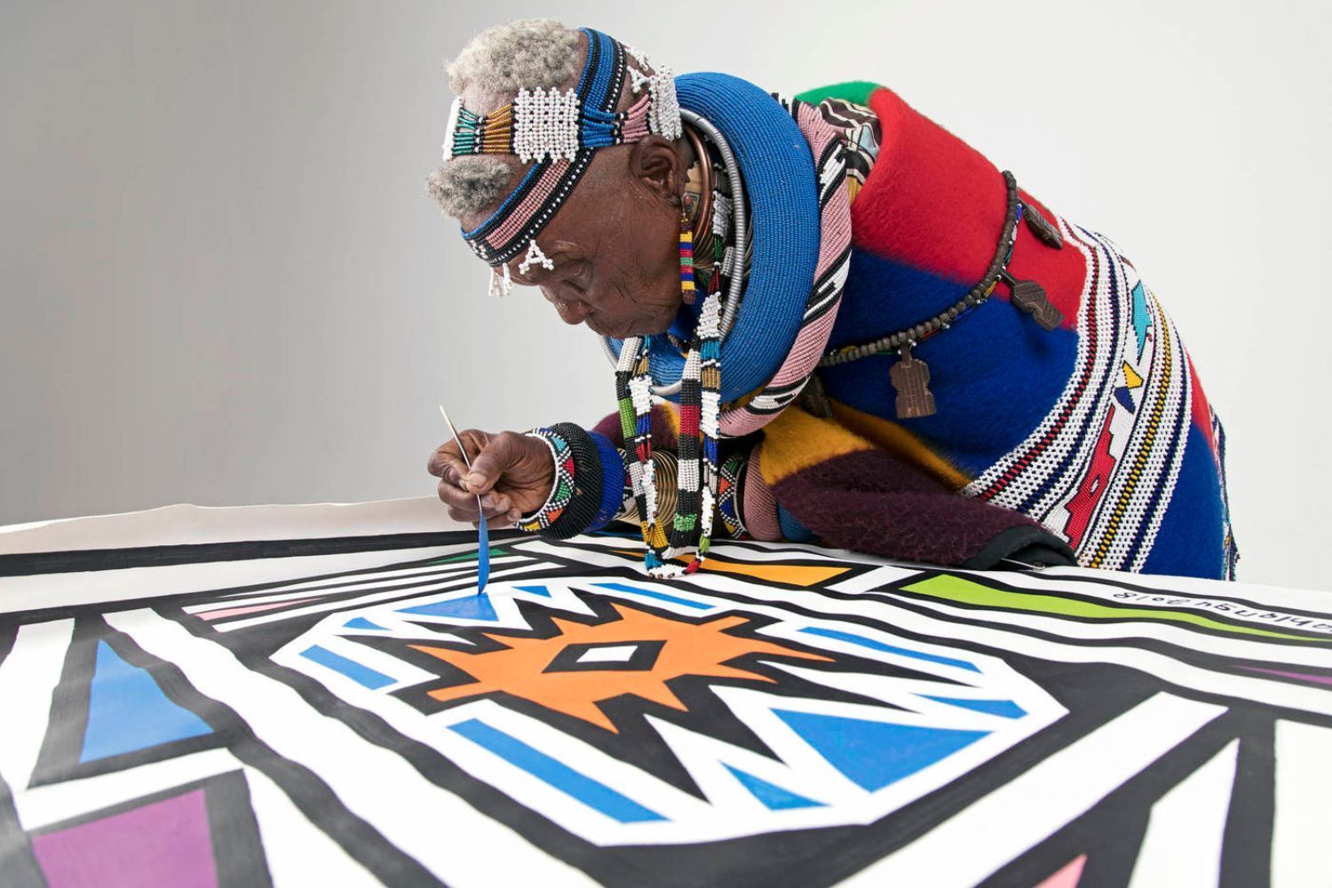 Iconic South African artist Esther Mahlangu, adorned in traditional Ndebele attire and intricate beadwork, leans over a large canvas as she carefully paints a vivid geometric pattern in blue, orange, black, and white. Her silver hair is styled in a crown-like shape and decorated with beads, and she wears colorful layers of fabric and jewelry, embodying the rich heritage of her culture.