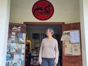 Karam Youssef, wearing a grey pullover, is standing in the entrance of her bookshop. Above her head, a red sign reads “Al Kotob Khan” in Arabic.