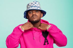 Kabza De Small wears a pink AMI Paris Ami de Coeur wool cardigan. He is also wearing an off-white and blue LV bucket hat, and is holding the collar of his sweater.