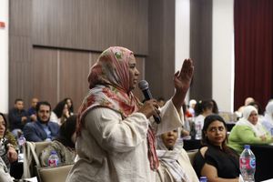 A woman wearing a beige and pink hijab is addressing people in a conference room, speaking into a microphone and gesticulating with a lifted arm.