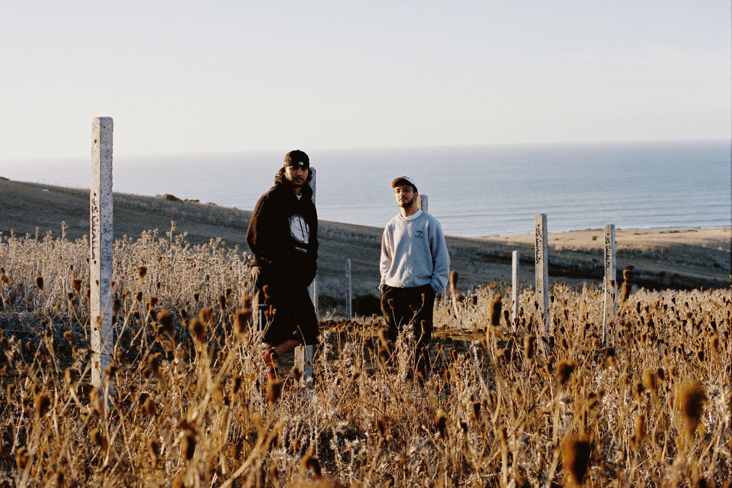 Small X and Saib standing in a field above the Atlantic Ocean, leaning on wooden poles with white paint. Small X is wearing a black jumper and cap, Saib a grey jumper and cap. 