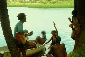 Tommy WÁ is photographed among friends. He is holding the guitar and looking out at the lake, while one woman sits in a boat.