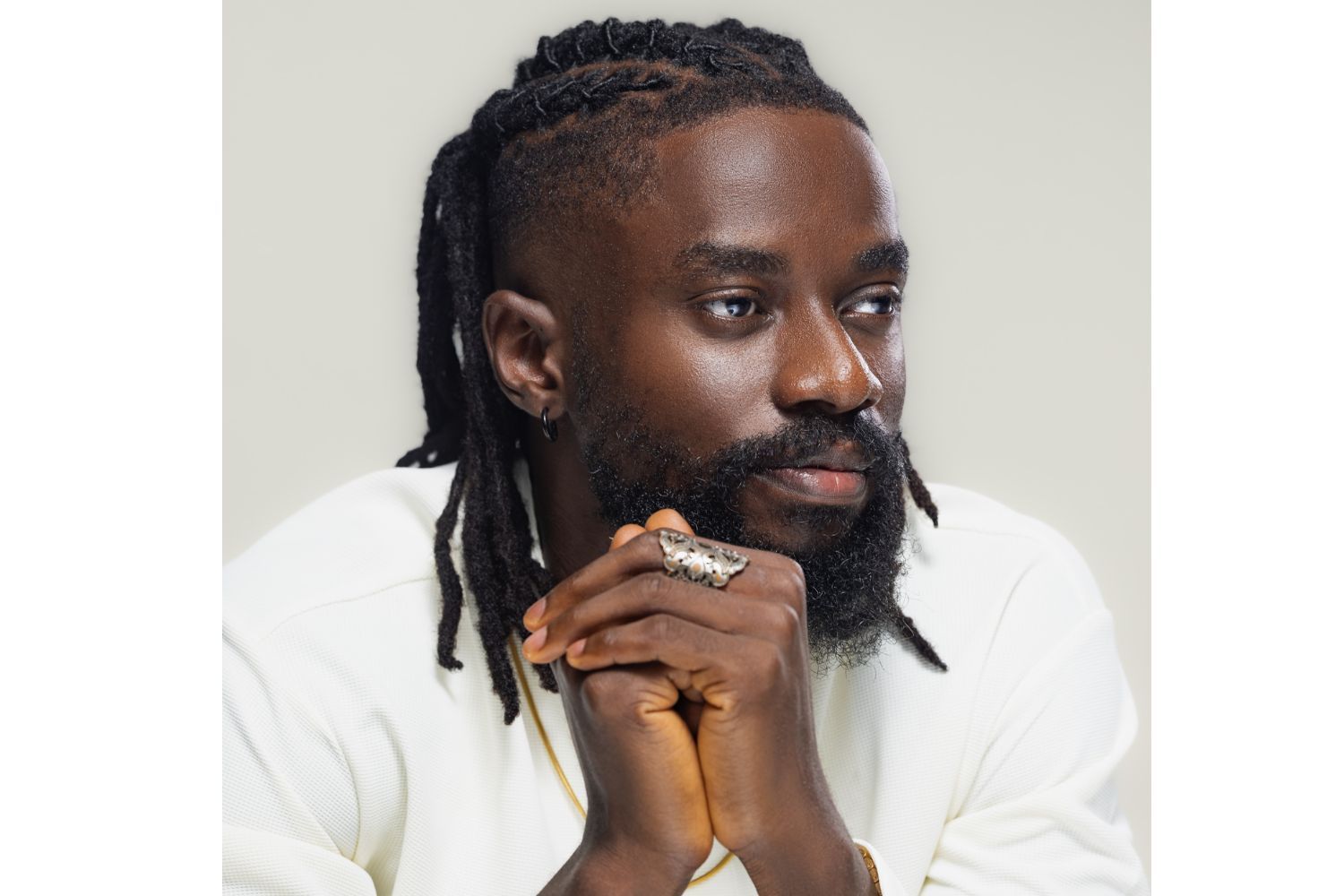A man with a full beard and long locks styled in cornrows is pictured in a close-up portrait. He’s wearing a white shirt and a silver statement ring shaped like a panther. His hands are clasped together near his face, and he gazes thoughtfully into the distance. The background is a soft, neutral grey, keeping the focus on his expression.