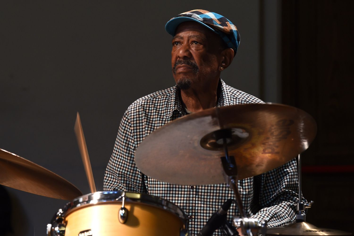 Louis Tebogo Moholo wears a checkered shirt and patterned cap and plays the drums, focused under soft stage lighting.
