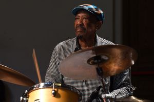 Louis Tebogo Moholo wears a checkered shirt and patterned cap and plays the drums, focused under soft stage lighting.