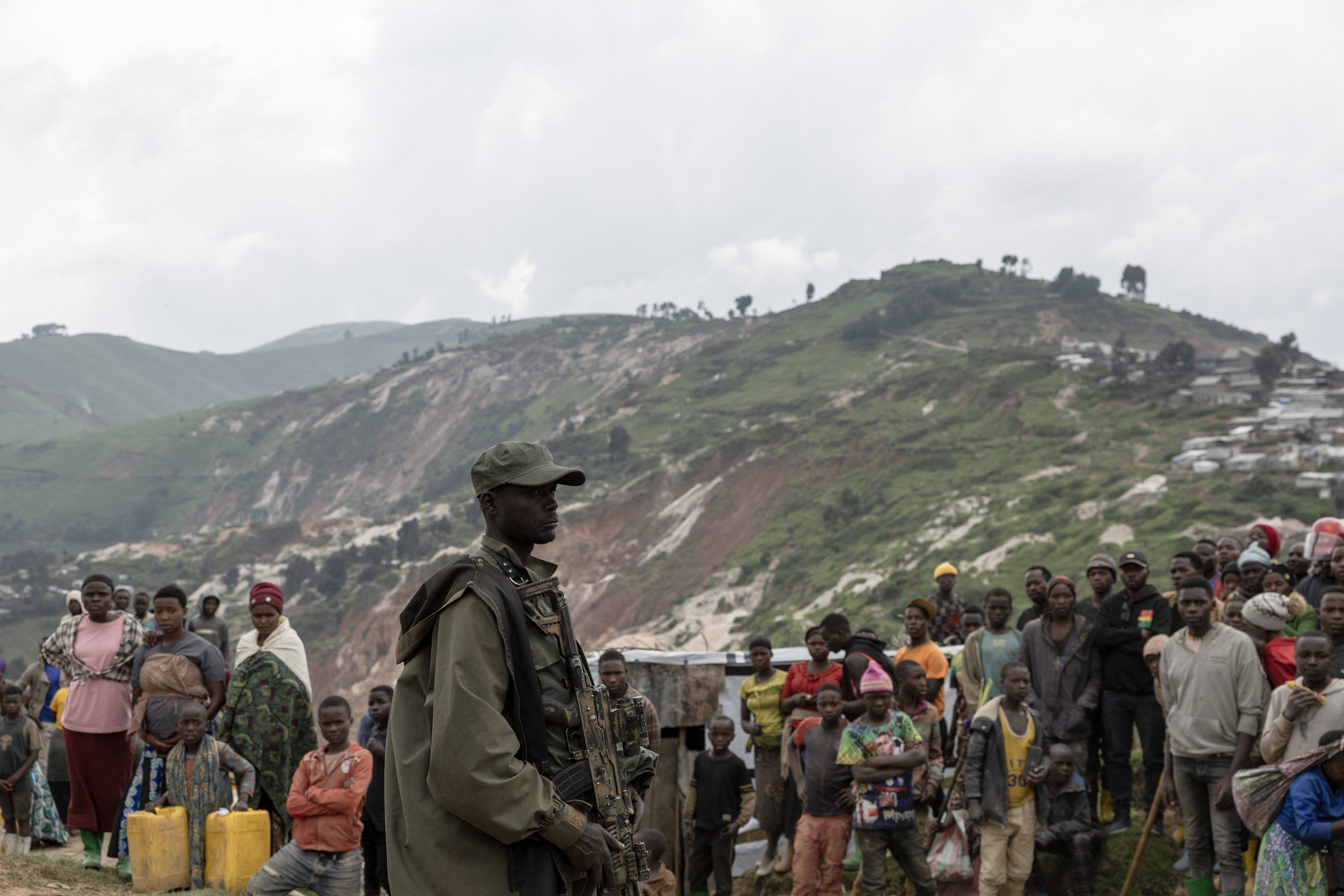 An armed M23 soldier stands at the Coltan mining pits in Rubaya on March 5, 2025. A group of people, adults and children, look on.