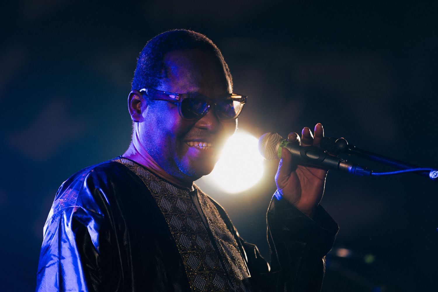 A close-up of Malian singer Amadou Bagayoko singing onstage, drenched in a blue light.