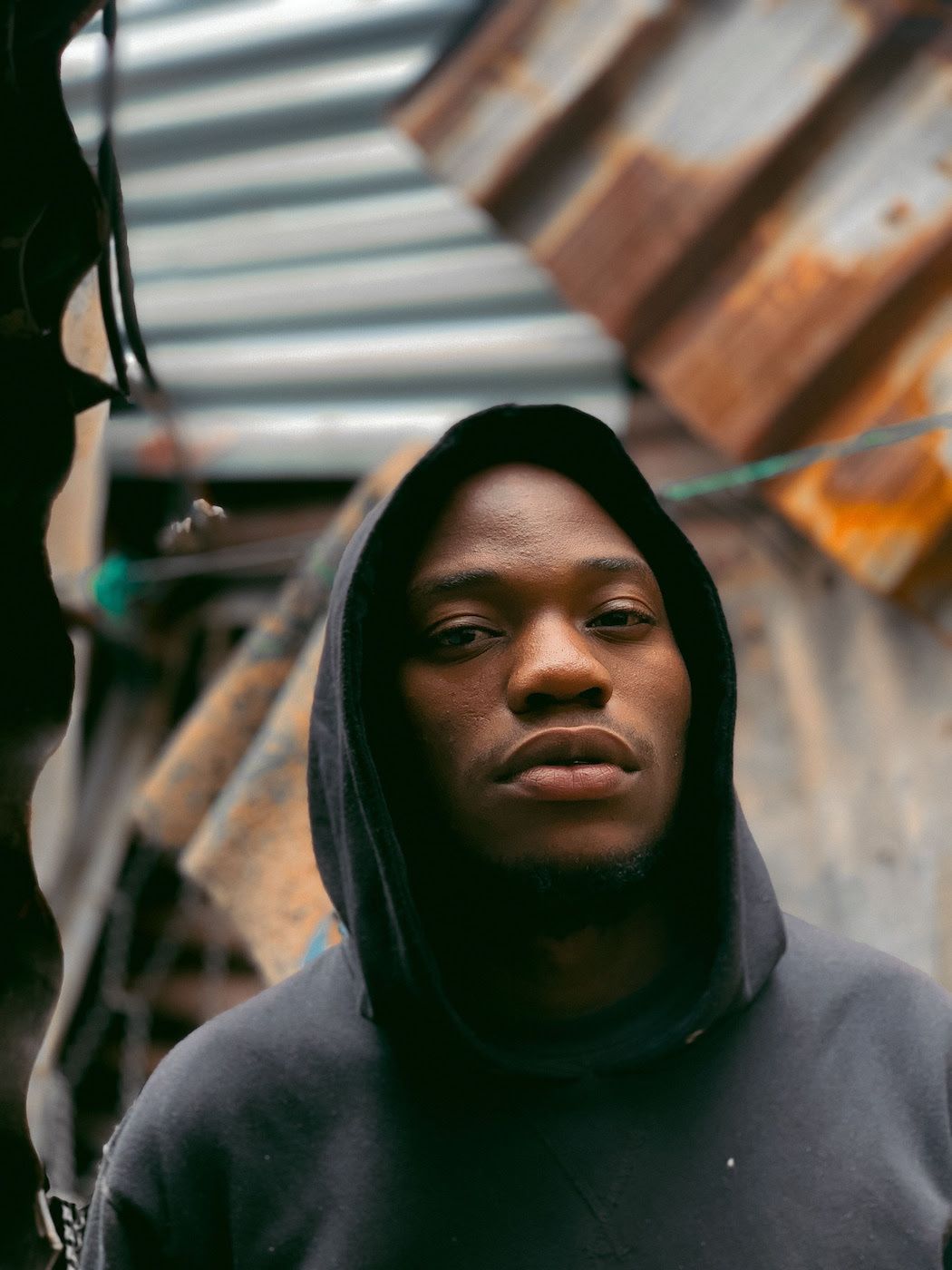 A close-up portrait of Marcus Garvey in a black hoodie standing among shacks. 