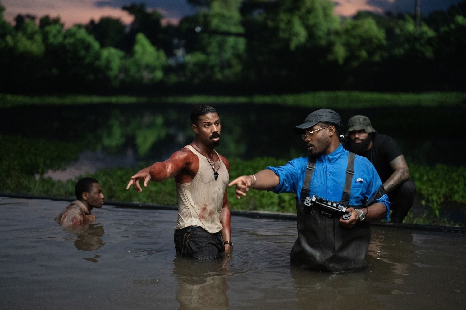 The actor Michael B. Jordan, wearing a wet vest stained with blood, is in a lake looking to the side, and pointing alongside director Ryan Coogler who is also pointing in the same direction, and wearing waders.  