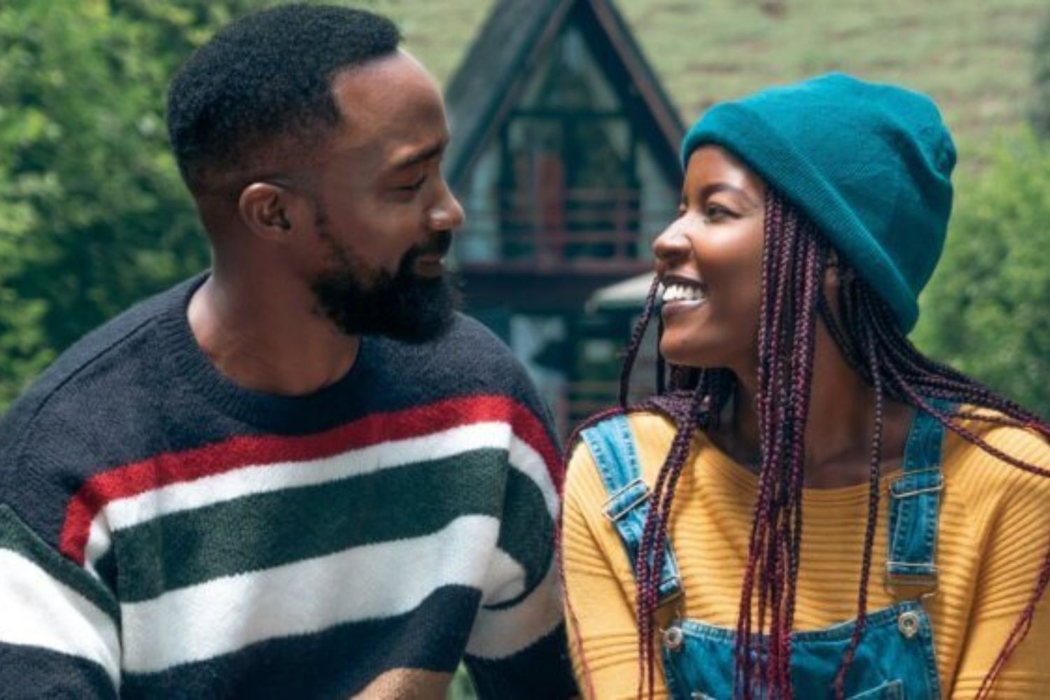 A man and woman, in a scene from the Kenyan film Sayari, sit close together outdoors, looking into each other’s eyes; the man wears a striped sweater, and the woman wears a teal beanie, mustard top, and denim overalls, with a cozy cabin and greenery in the background.