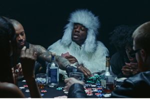 A group of people sit around a poker table covered in chips and drinks, with one man in the center wearing a dramatic white fur coat and hat, commanding attention with his bold outfit and relaxed demeanor.