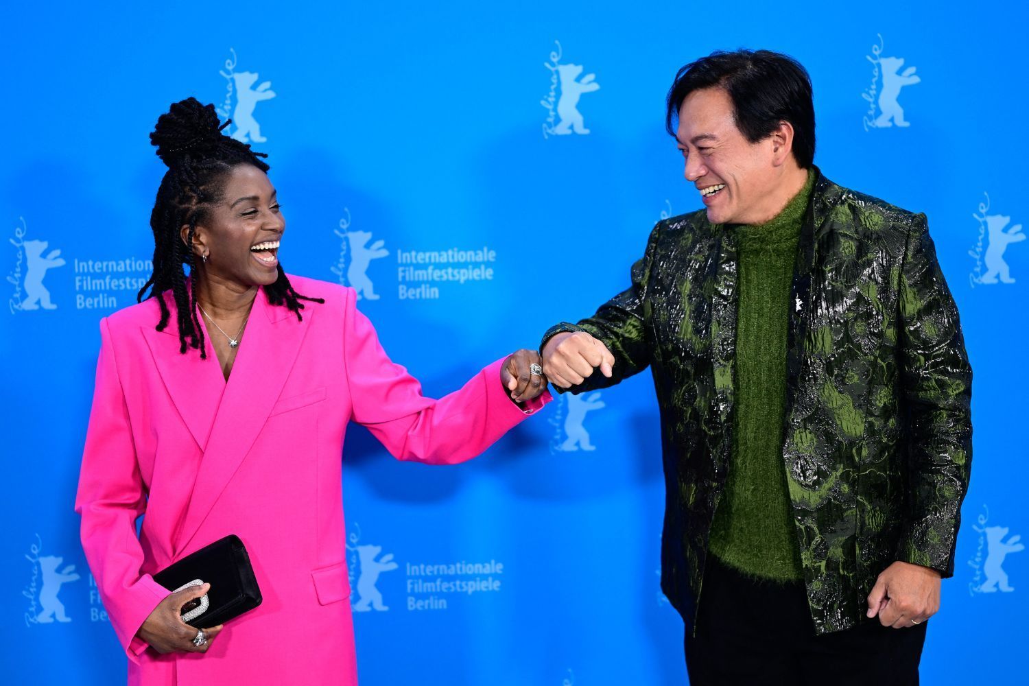 A smiling Black woman with long locks styled in a top bun, wearing a bright pink suit, fist-bumps an Asian man in a textured green suit and sweater; they stand in front of a blue backdrop for the Berlin International Film Festival, sharing a joyful moment.