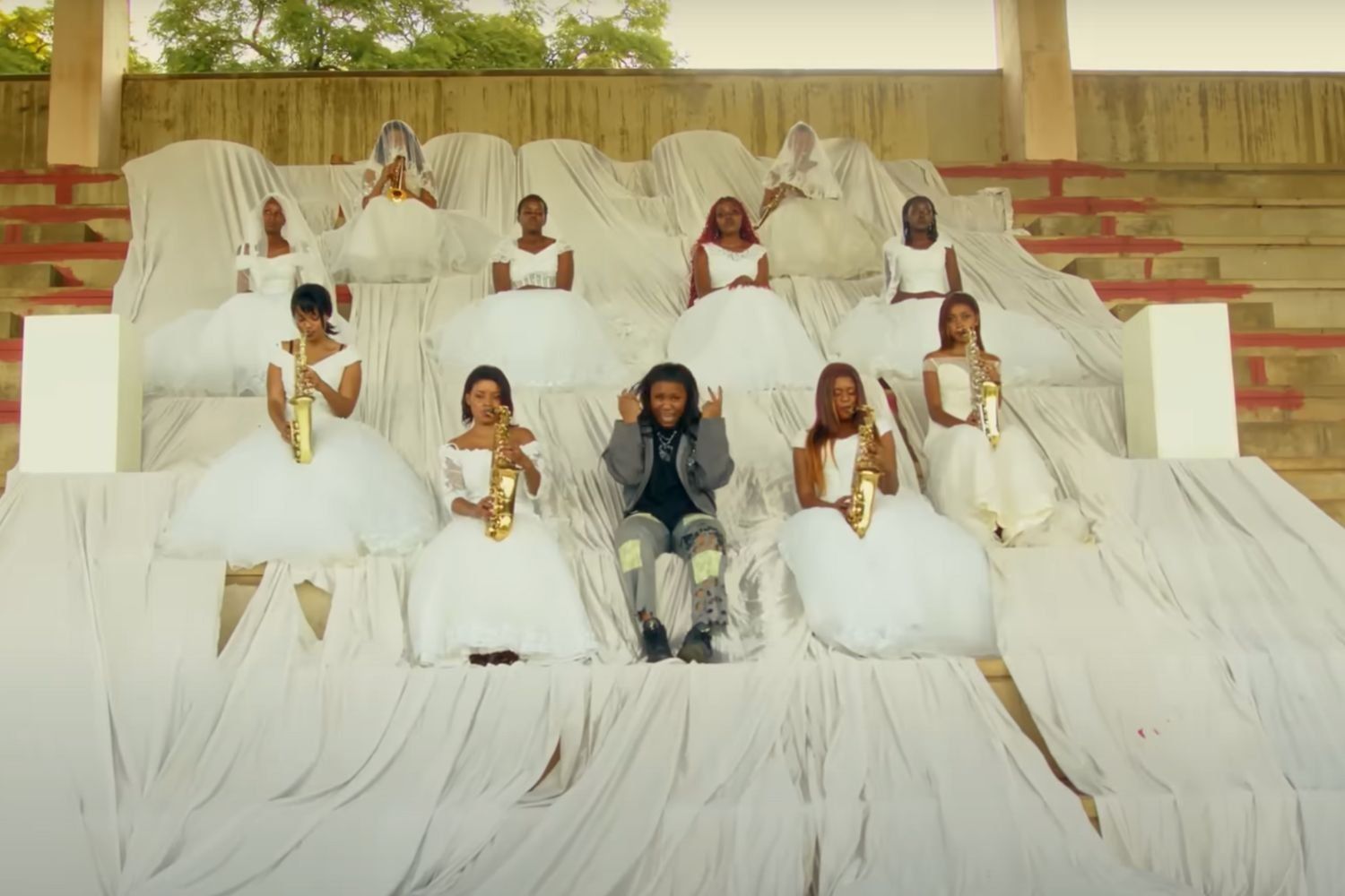 Zimbabwean artist Nisha Ts performs alongside an all-female band of saxophone players. They sit on stairs draped with white cloth. The saxophone players are wearing white gowns. Nisha Ts wears denim.