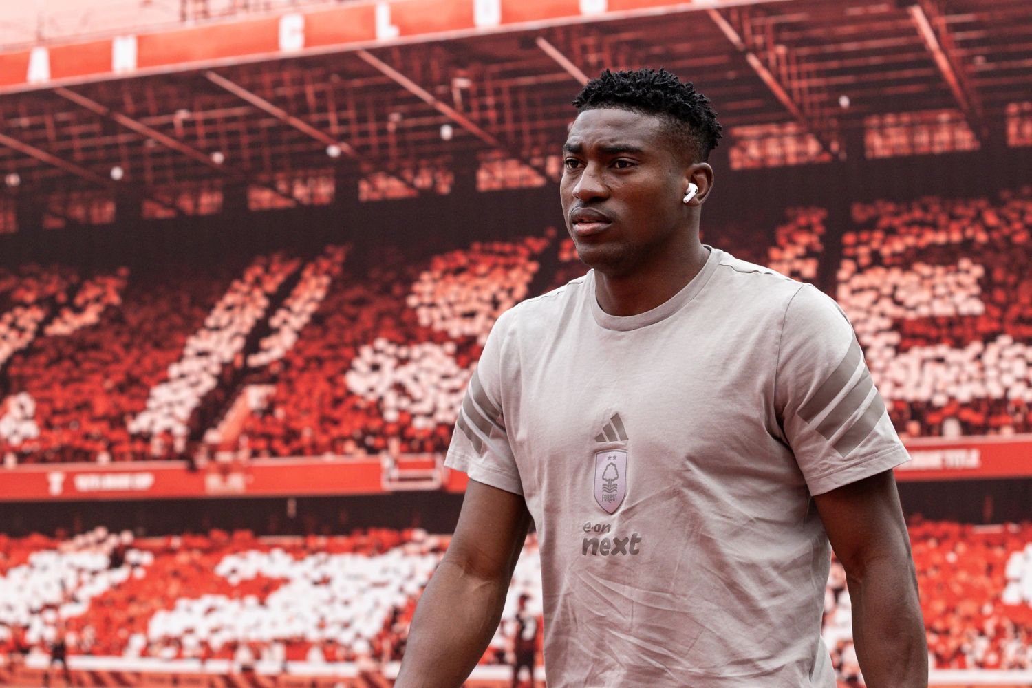 Taiwo Awoniyi walks across a stadium pitch, wearing a grey Nottingham Forest t-shirt and wireless earbuds. Behind him the stands are filled with red and white fan cards forming a coordinated display. 