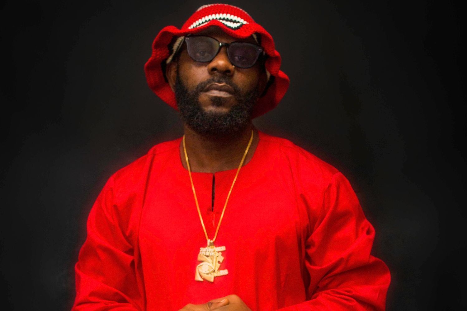 ODUMODUBLVCK poses for the camera against a dark background, wearing a bright red outfit and a matching red knit bucket hat; he has a full beard, glasses, and a gold necklace with a large pendant.