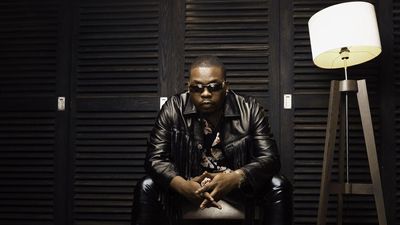 Nigerian star Olamide sits in a dark room dressed in all black leather and snake skin cowboy boots. 