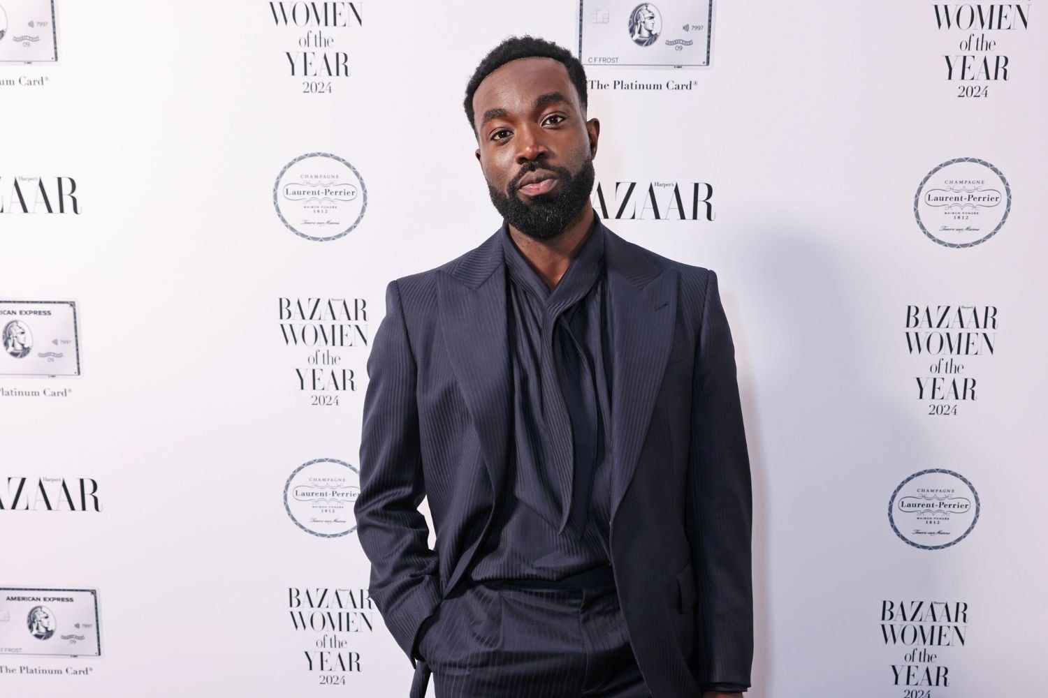 Paapa Essiedu poses on a red carpet at the “Harper’s Bazaar Women of the Year 2024” event. He is dressed in a dark, pinstriped suit with a relaxed fit and a stylish draped shirt underneath. The backdrop features logos from Harper’s Bazaar, American Express, and Laurent-Perrier, among others.