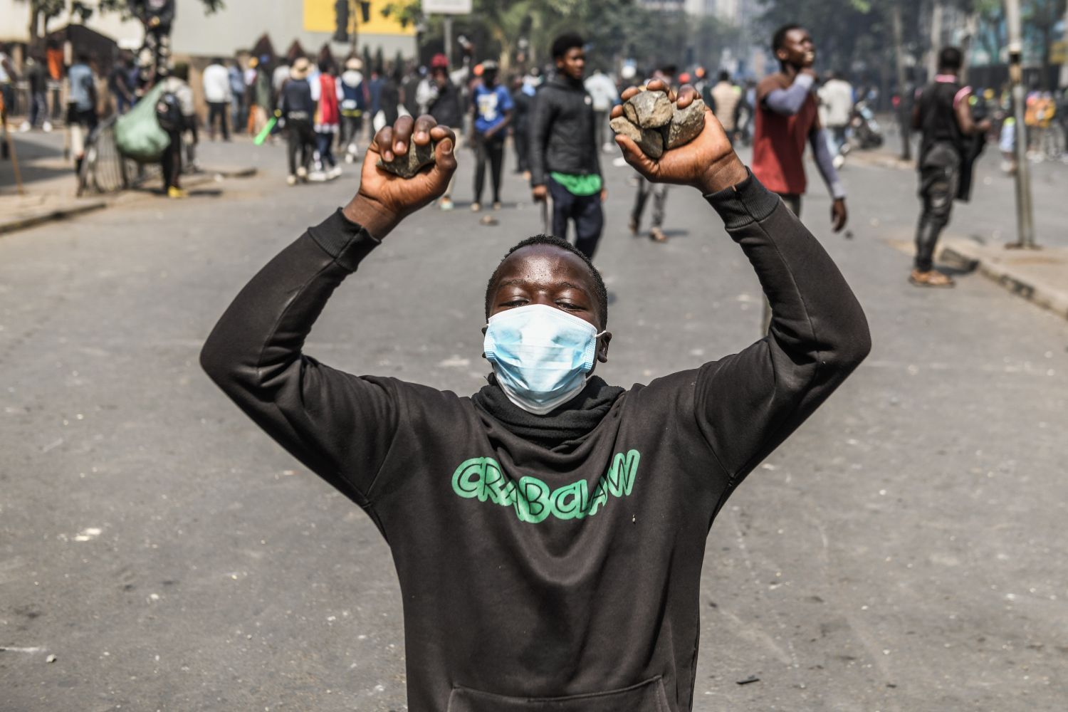 A young man with a mask partially covering his face kneels with his hands up, holding rocks, during protests against police brutality at the Central Business District in Nairobi, Kenya.