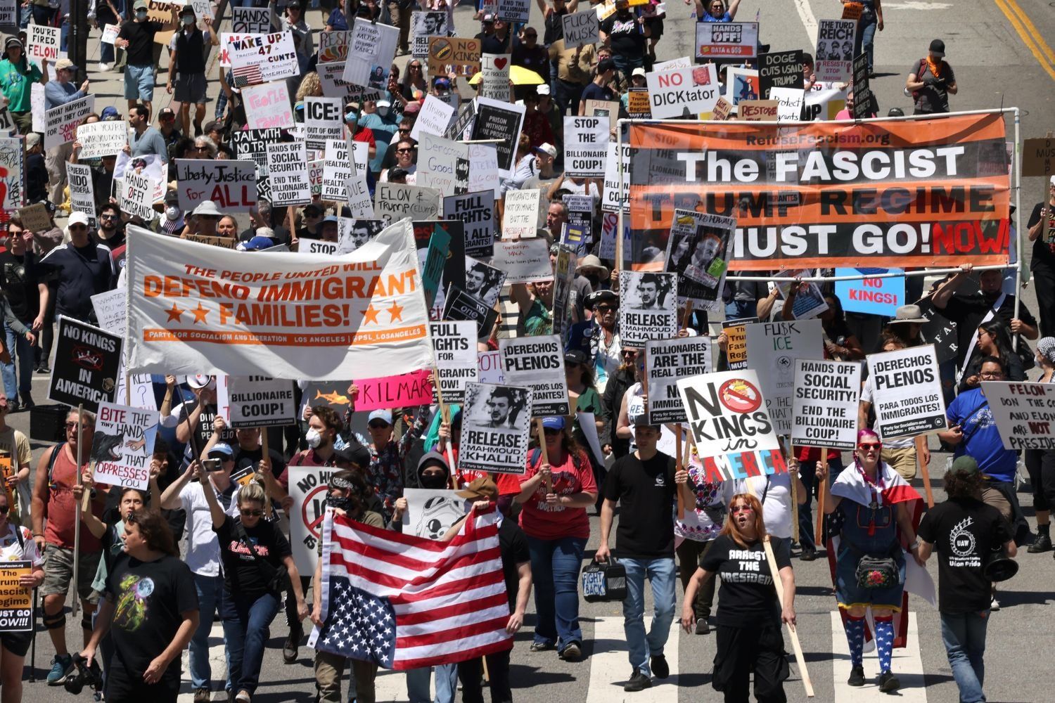 A large crowd protests in the street, holding signs demanding immigrant rights, social justice, and the end of the Trump regime.