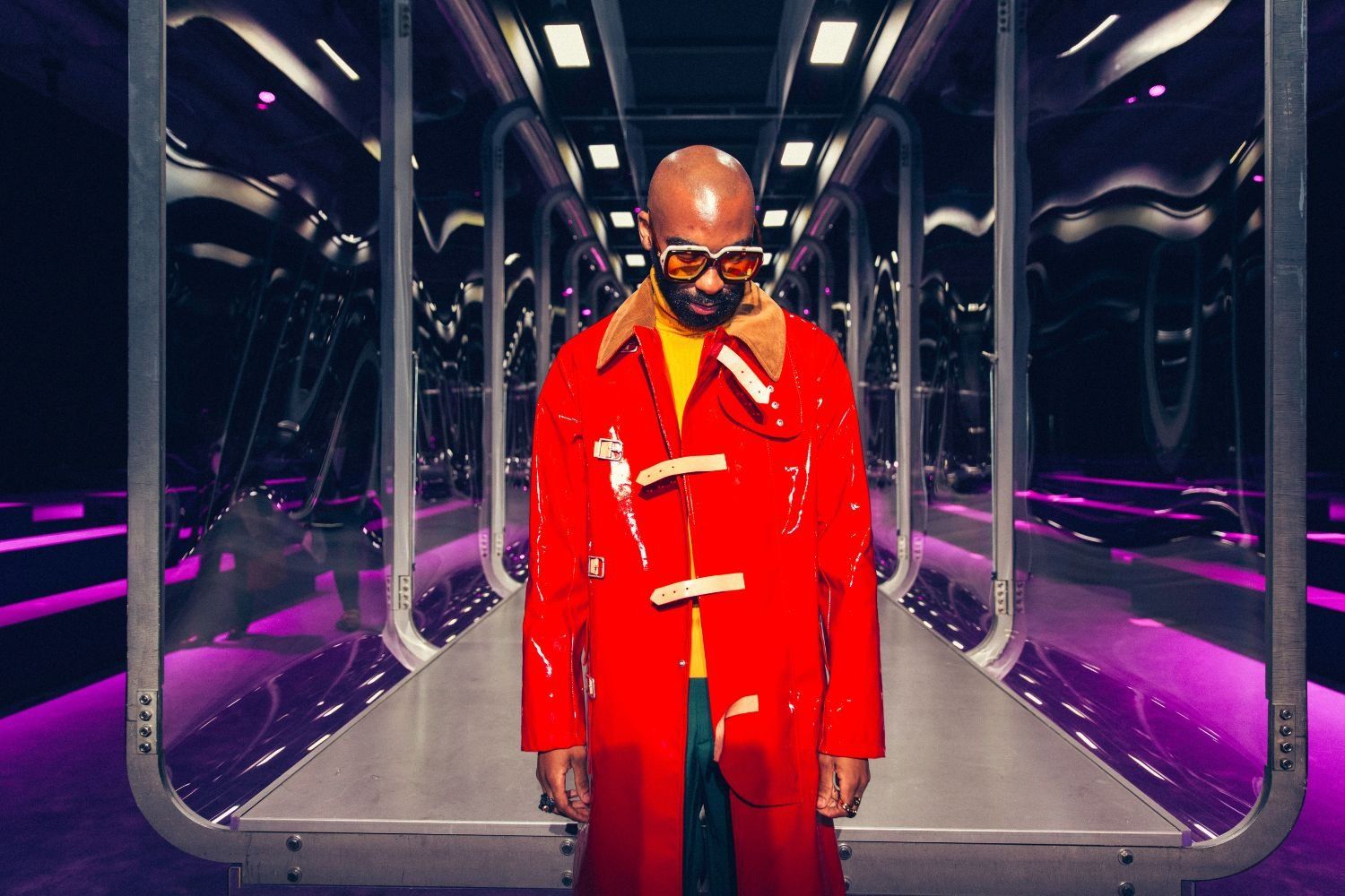 South African rapper, the late Riky Rick, poses for the camera. He is bald, wearing a shiny red coat and large sunglasses standing on a futuristic walkway lit with purple lights, looking down.