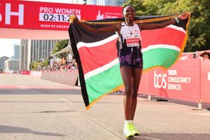 Ruth Chepngetich smiles and holds the Kenyan flag on her shoulders, with a marathon finish line behind her. 
