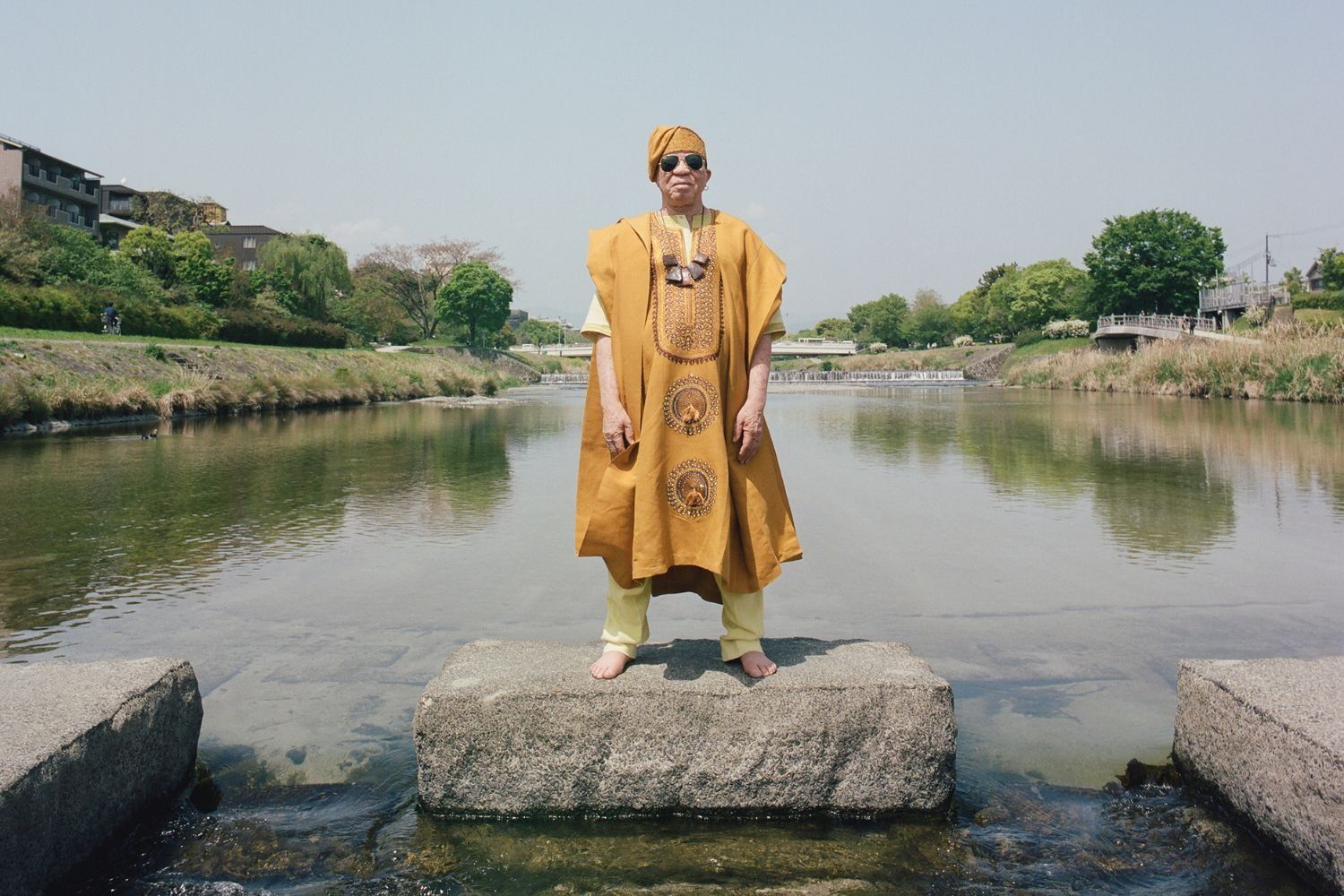 Salif Keita, wearing a mustard yellow traditional outfit and sunglasses stands barefoot on a stone block in a shallow river, with greenery, buildings, and a clear sky in the background.