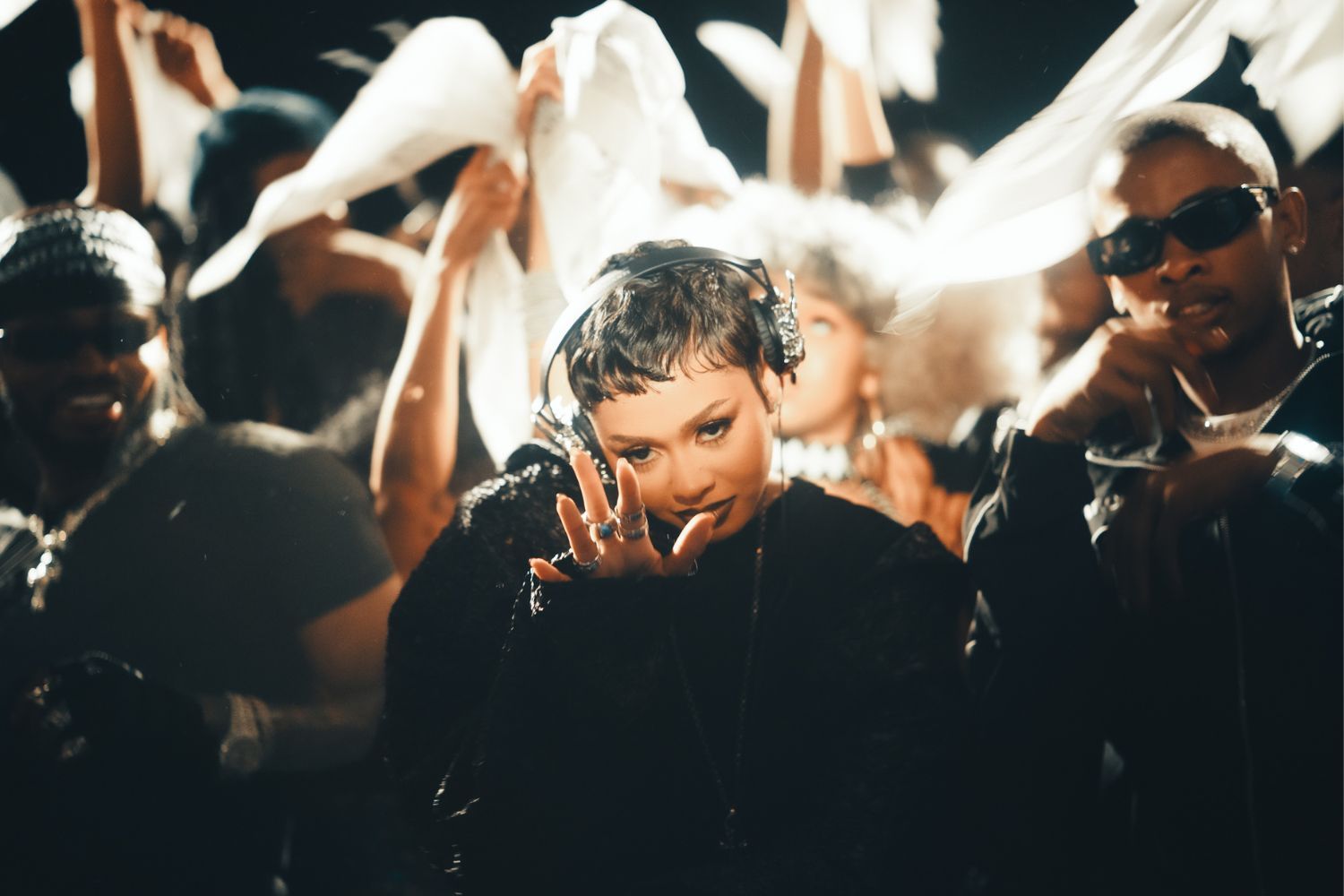 Still from Skyla Tylaa’s “Bombshell (Cheza)” video. She stands in the spotlight wearing headphones and dark clothing, surrounded by a lively crowd waving white cloths and dancing under dramatic lighting. 