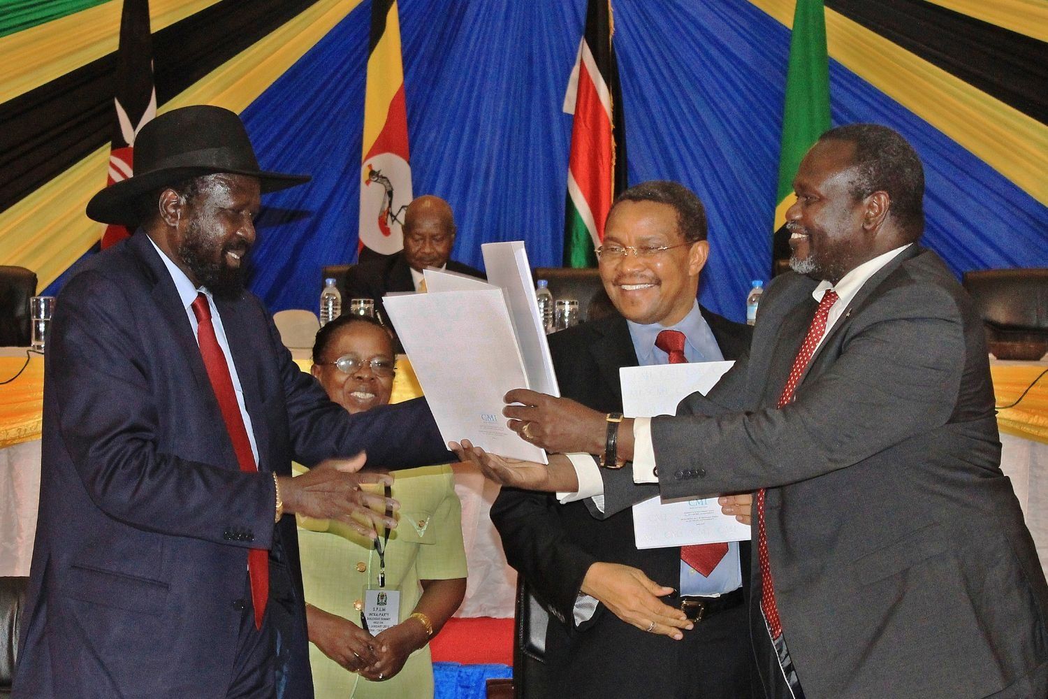 South Sudanese President Salva Kiir, wearing a dark suit, red tie, and wide-brimmed black hat, shakes hands and exchanges documents with opposition leader Riek Machar, also in a dark suit and red tie, as they smile during a peace agreement ceremony. Former Tanzanian President Jakaya Kikwete stands between them, grinning and holding papers, while other officials and colorful East African flags form the backdrop.