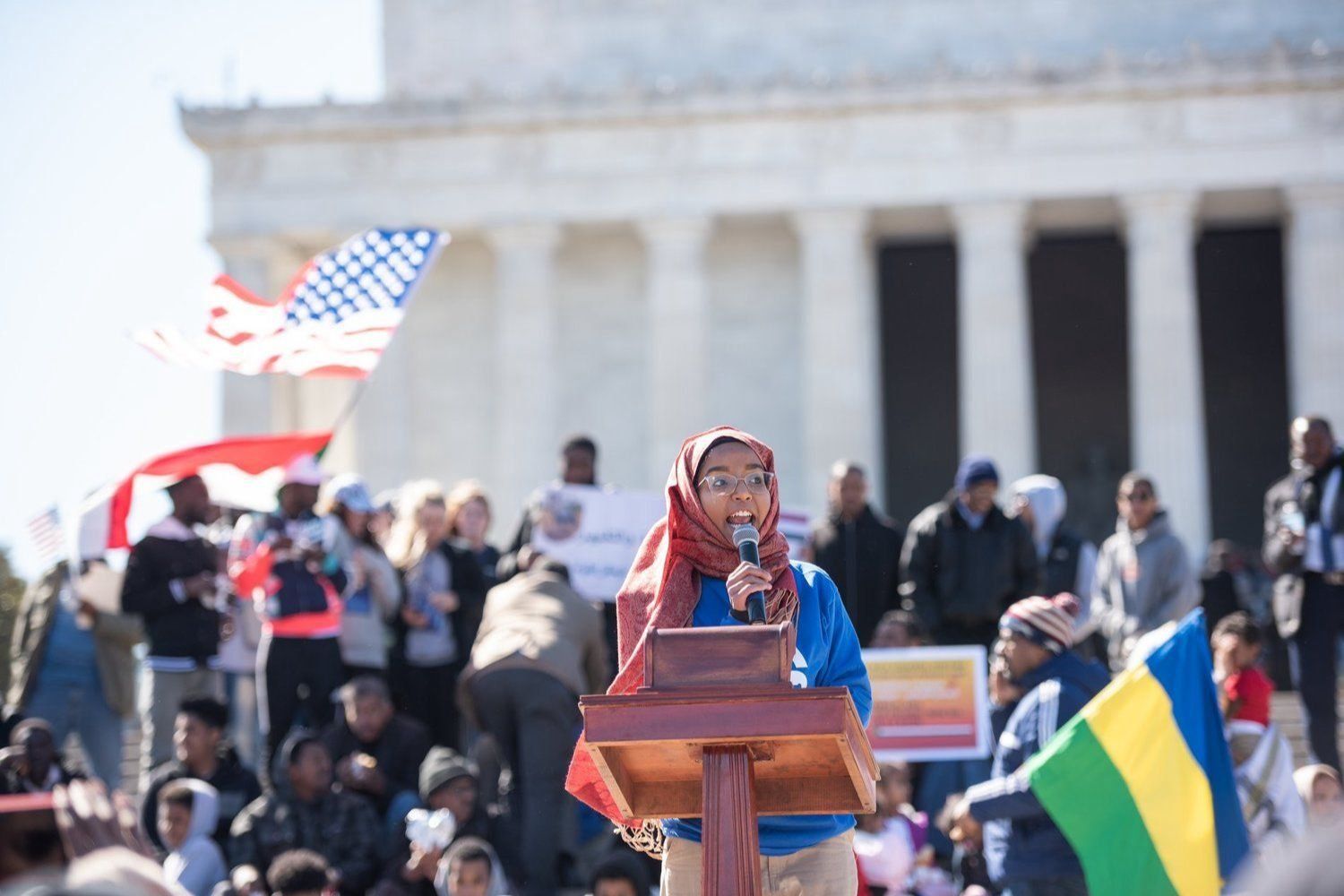 Sudanese American activist and poet speaking at a rally in Washington D.C., protesters are holding Sudanese and American flags behind her. 