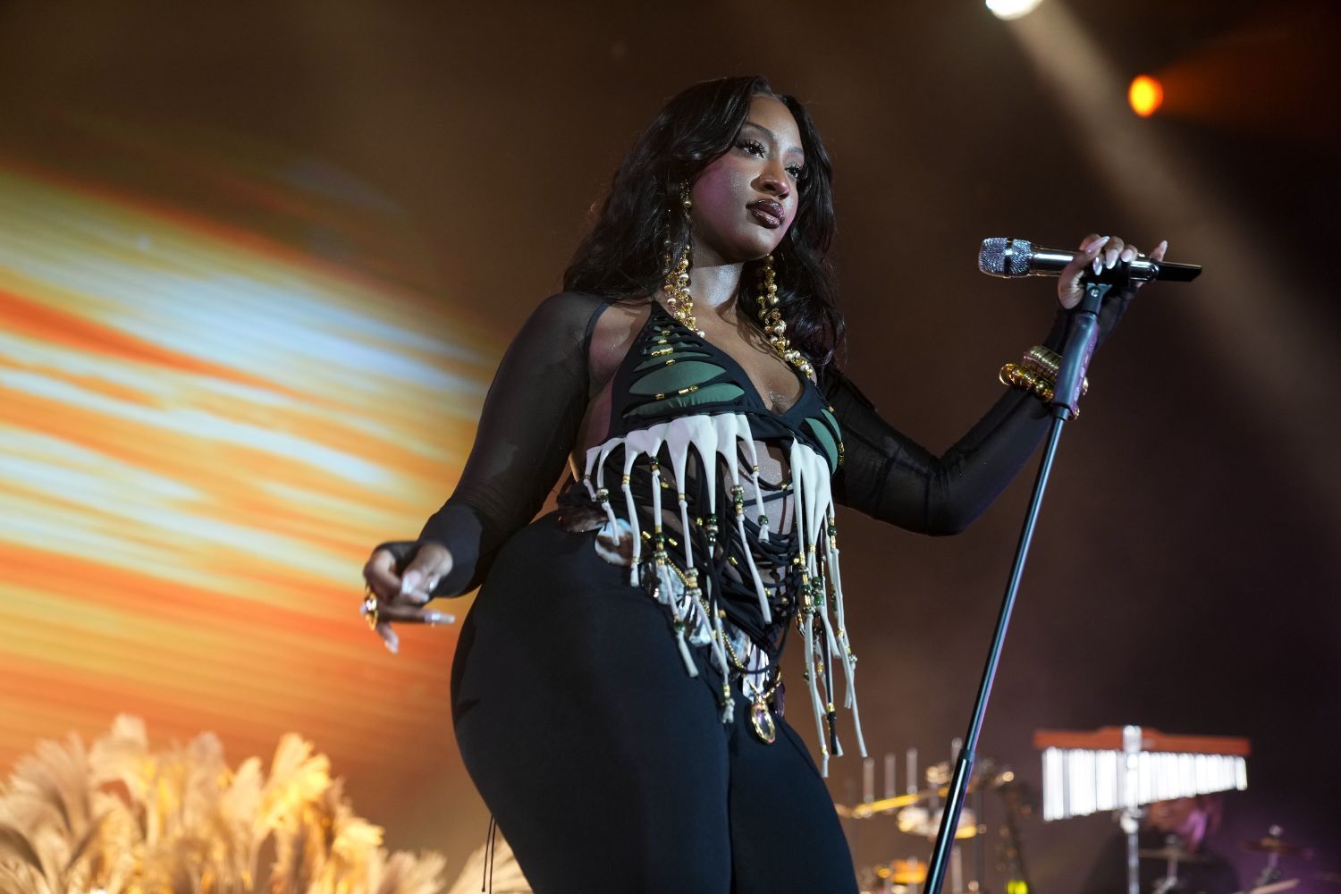 Tems in a bold stage outfit sings into a microphone during a live concert under dramatic lighting.
