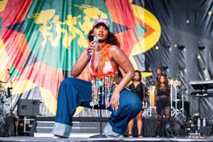 Tems kneels on stage mid-performance, holding a microphone. She wears a colorful halter top with beaded fringe, wide-leg jeans, and a pink cap. A band and backup singers dressed in black stand behind her, with a bold African-themed stage backdrop in red, yellow, green, and black.